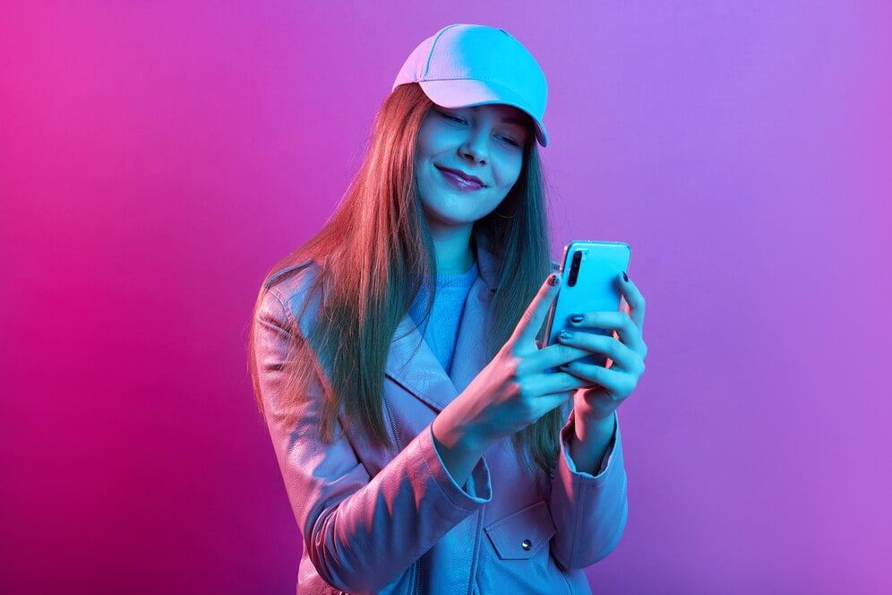 Retrato de uma jovem e bela modelo elegante usando couro padeiro e boné de beisebol, segurando o telefone inteligente nas mãos
