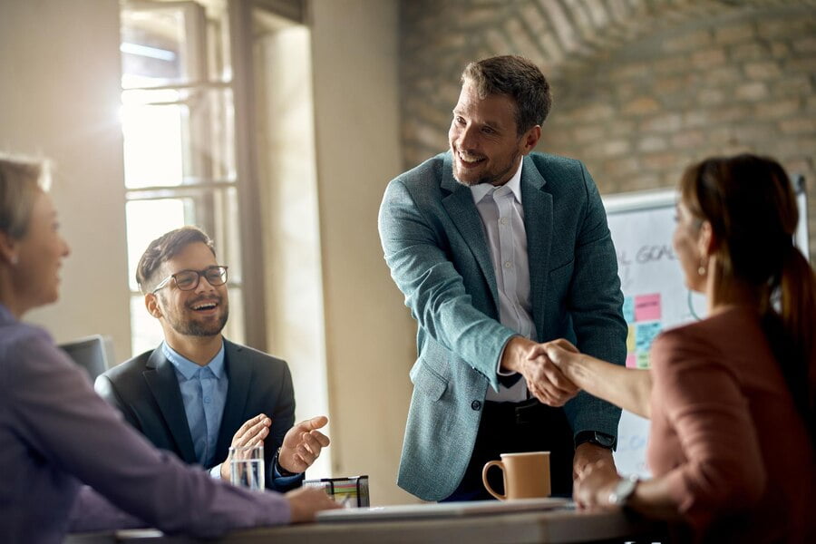 5 Tendências de Marketing B2B para Conquistar Novos Clientes 1 Empresários felizes cumprimentando seus colegas em uma reunião e apertando as mãos de um deles no escritório