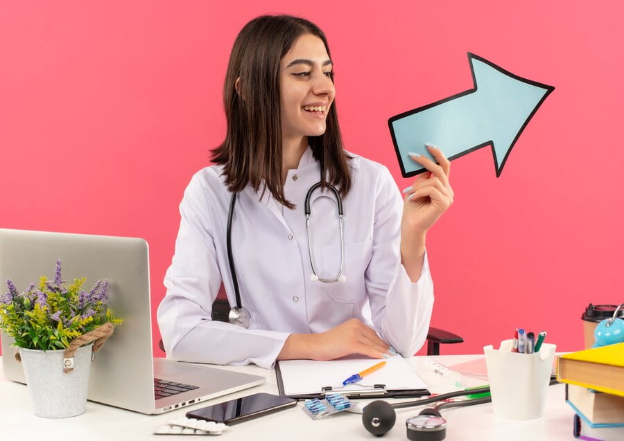Health Marketing: Saúde e Bem-estar em Destaque 1 Jovem médica de jaleco branco com estetoscópio no pescoço segurando uma seta azul olhando de lado sorrindo sentado à mesa com o laptop sobre a parede rosa