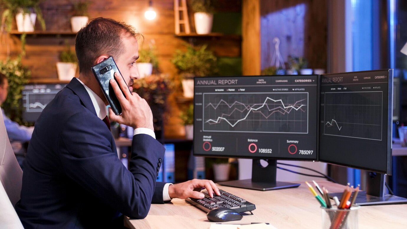 O gerente fala ao telefone enquanto analisa alguns gráficos no monitor. Duas telas