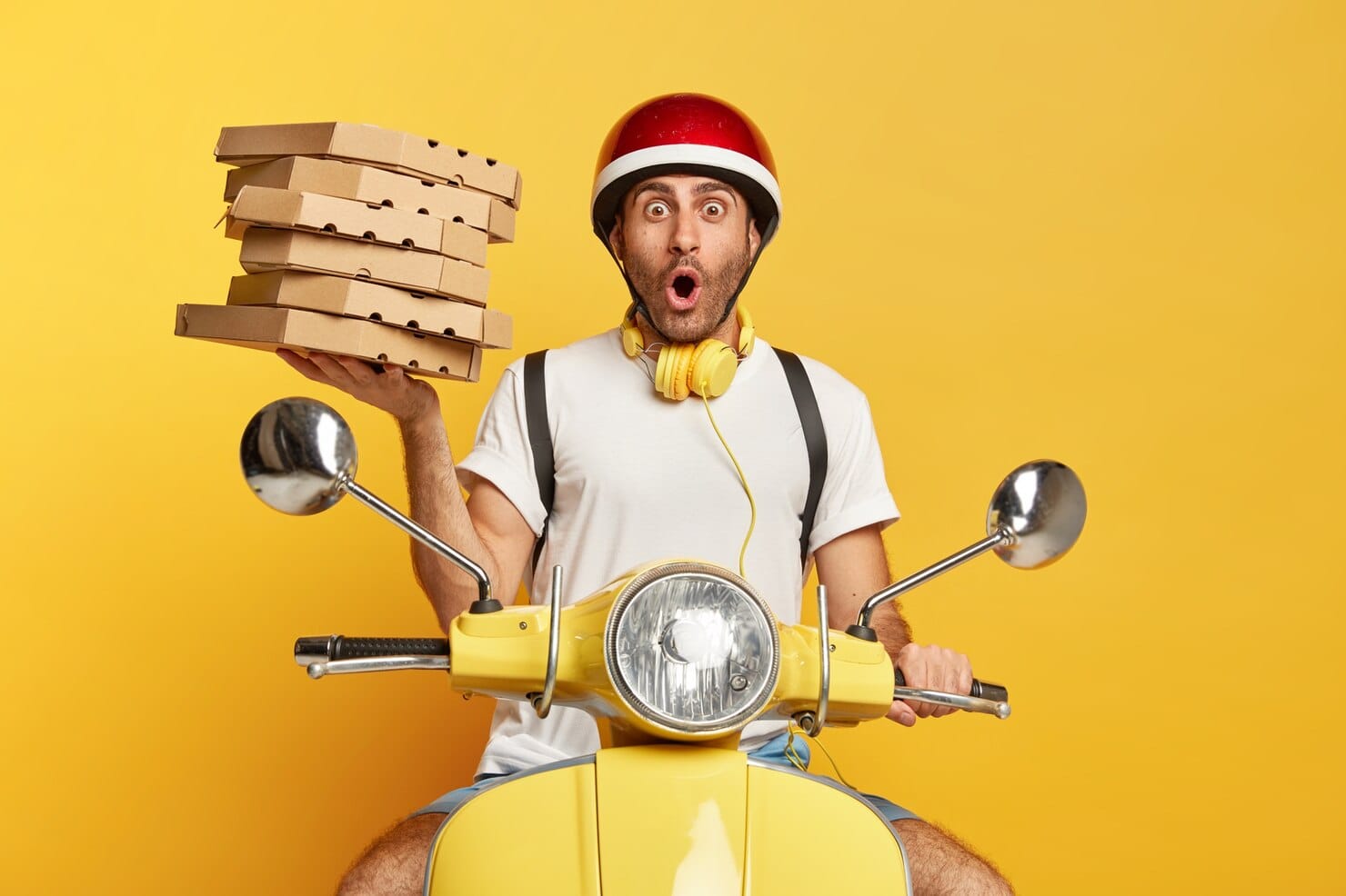 Entregador chocado com capacete dirigindo uma scooter amarela enquanto segura caixas de pizza