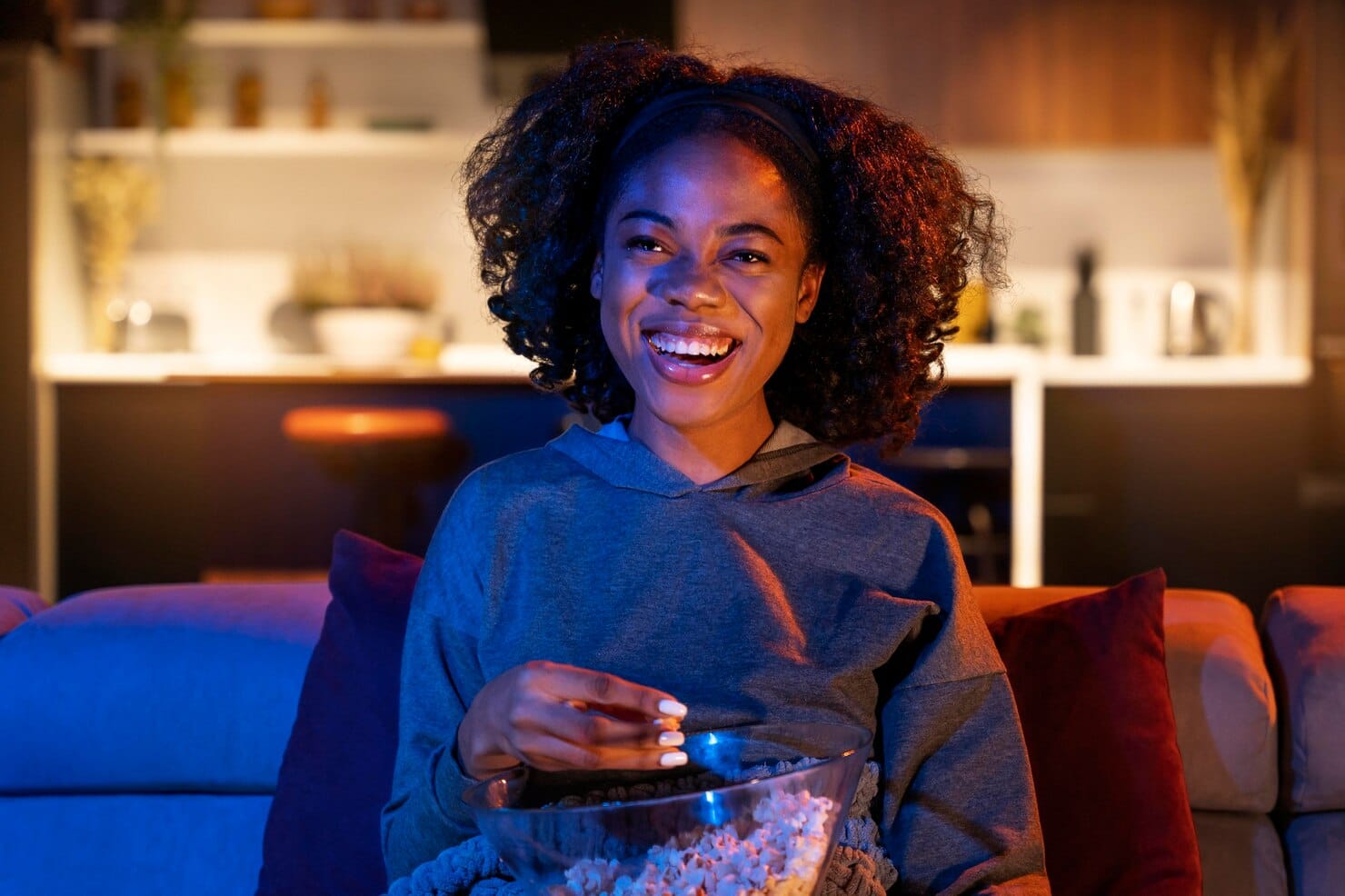 Mulher sorrindo enquanto assiste TV no sofá e come pipoca em ambiente aconchegante.