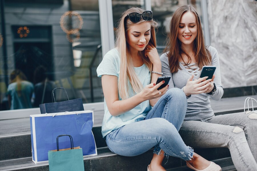 Duas Mulheres sentadas com sacola de compras mexendo no celular