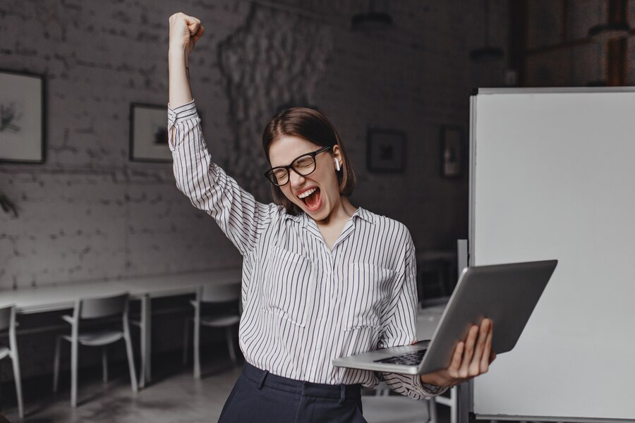 Mulher de negócios com o laptop na mão está feliz com o sucesso retrato de mulher de óculos e blusa listrada gritando com entusiasmo e fazendo gesto vencedor