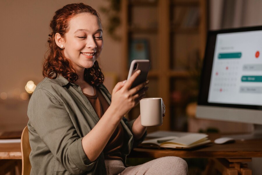 Mulher em casa usando smartphone na frente do computador enquanto toma café