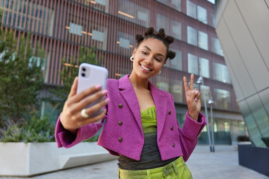 Garota atraente com roupa elegante cria conteúdo de influência mostra o símbolo da paz em poses de câmera frontal do smartphone.