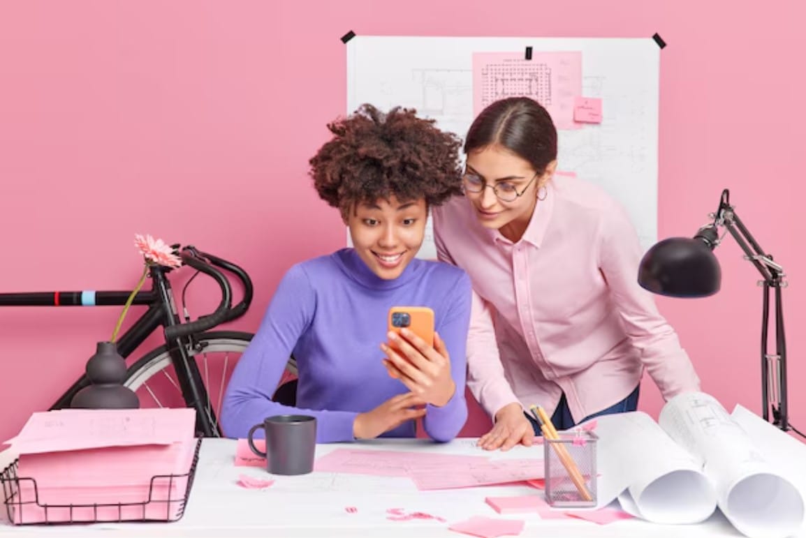 15 tendências de marketing para o segundo semestre de 2025 Conceito de cooperação e trabalho em equipe. Duas colegas de trabalho verificam o celular, trabalham nas plantas de uma nova casa ou na pose de um prédio no escritório no desktop