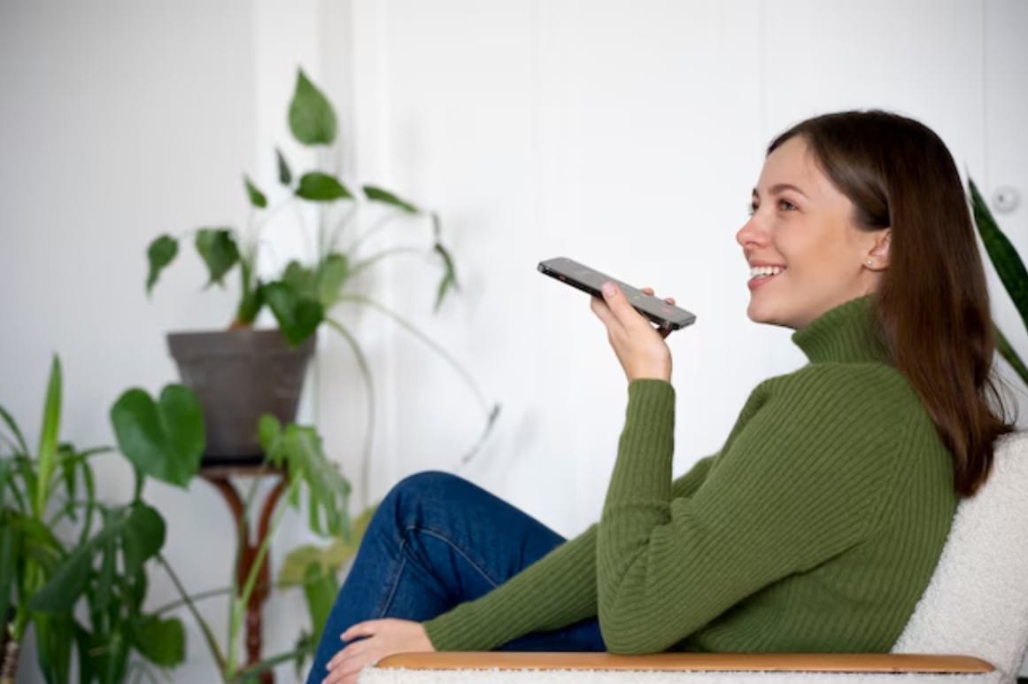 Pesquisa Por Voz no E-Commerce: Como Vender Mais? Mulher falando em seu smartphone enquanto está em casa usando o recurso viva-voz