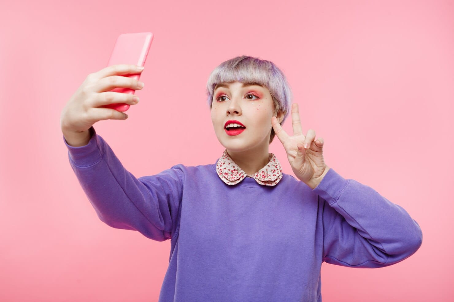 Como criar uma estratégia de publicidade com influenciadores virtuais Retrato do close-up da linda garota dollish com cabelo violeta curto curto, vestindo blusa lilás fazendo selfie sobre parede rosa