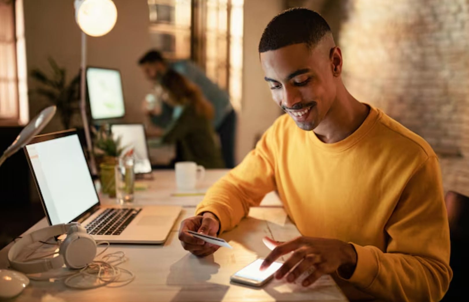 Feliz empresário afro-americano usando telefone celular e cartão de crédito de pagamentos online enquanto trabalhava à noite no escritório 