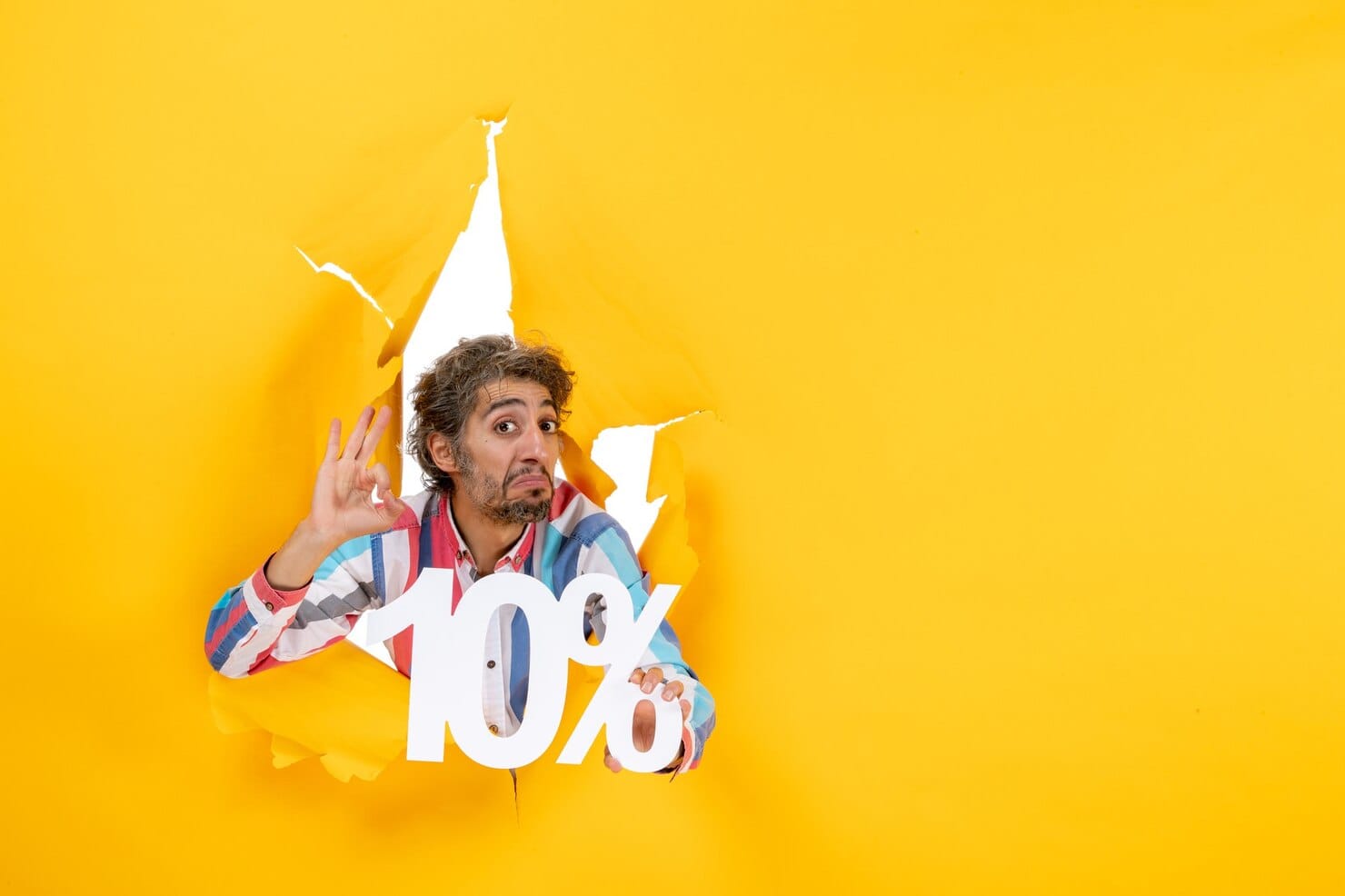 Vista frontal de um jovem surpreso mostrando dez por cento e fazendo gesto de óculos em um buraco rasgado em papel amarelo